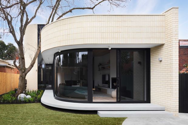 Exterior view of a contemporary residential extension with a distinctive curved white brick upper facade and rounded projecting form. Floor-to-ceiling dark-framed glazing opens onto a small stepped terrace, revealing a bright interior living area. The house sits beside a landscaped lawn with mature trees and timber fencing, with neighbouring brickwork visible at the sides.