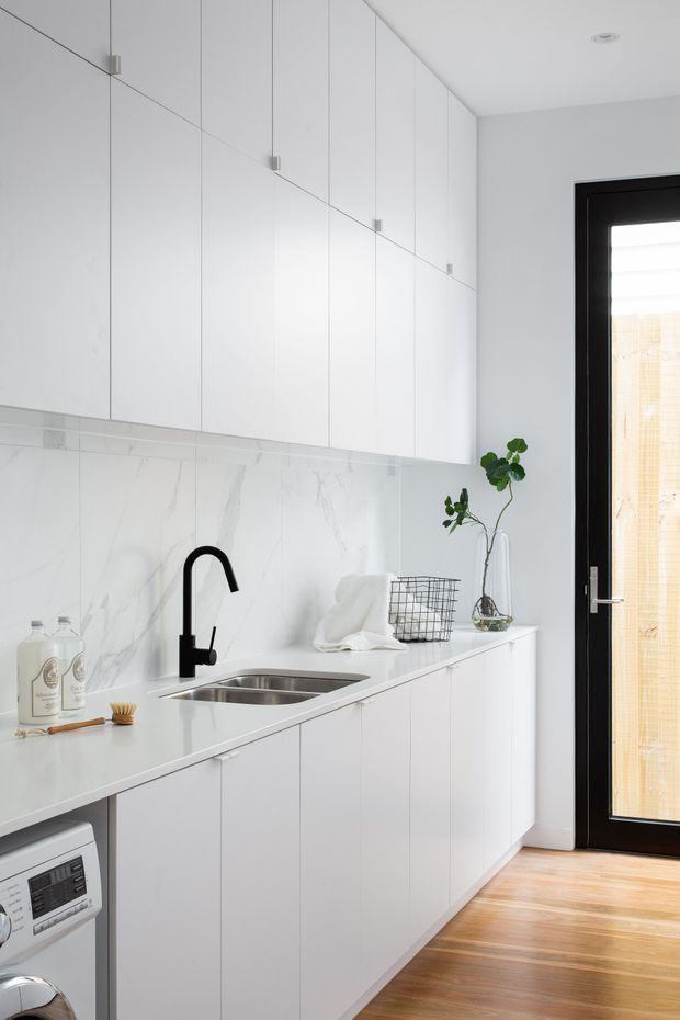 Bright, compact contemporary kitchen-laundry space with streamlined white handleless cabinetry, a white marble-look splashback and benchtop, black gooseneck tap, stainless-steel double sink, and warm timber flooring. A black-framed glazed door provides natural light, while minimal styling and indoor plants add subtle contrast.