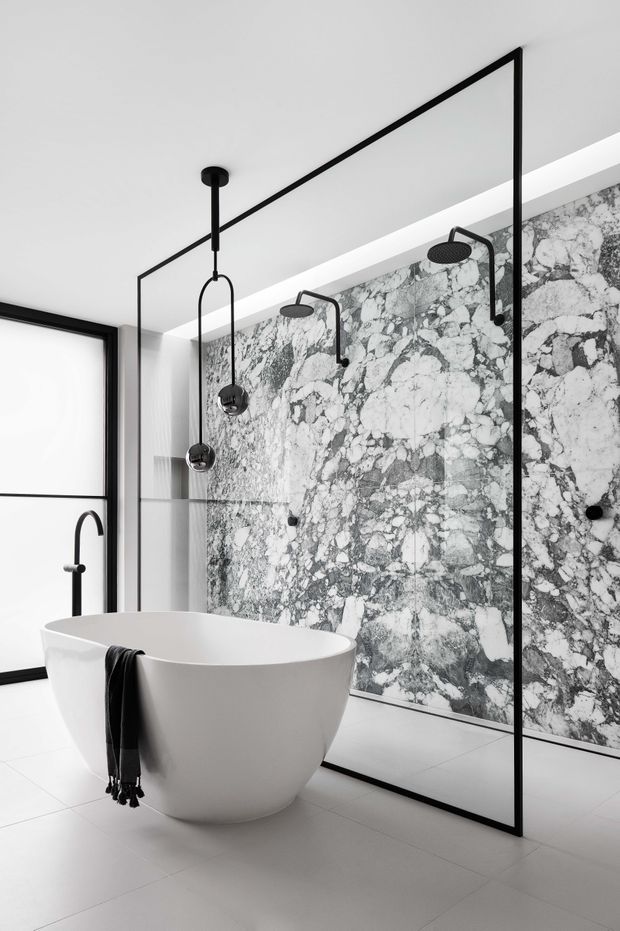 A high-end minimalist bathroom featuring a freestanding white bathtub, a frameless-style black metal shower enclosure, multiple matte black shower fixtures, large-format light floor tiles, and a dramatic grey-and-white veined stone feature wall. A large black-framed window provides natural light.