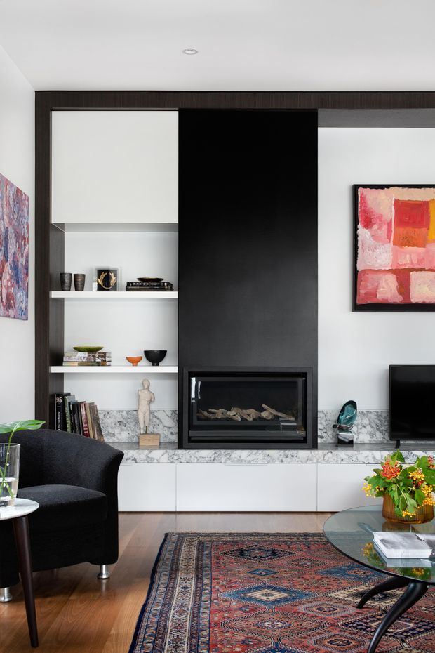 A refined living room centred on a built-in fireplace wall. Dark timber-toned framing surrounds open display shelving, a tall black fireplace surround, and white lower cabinetry with a grey-veined stone ledge. The room includes a black upholstered armchair, patterned area rug, low oval coffee table, television, colourful abstract artwork, books, and decorative objects. The overall look combines a monochromatic architectural backdrop with warm timber flooring and vivid art and textiles.