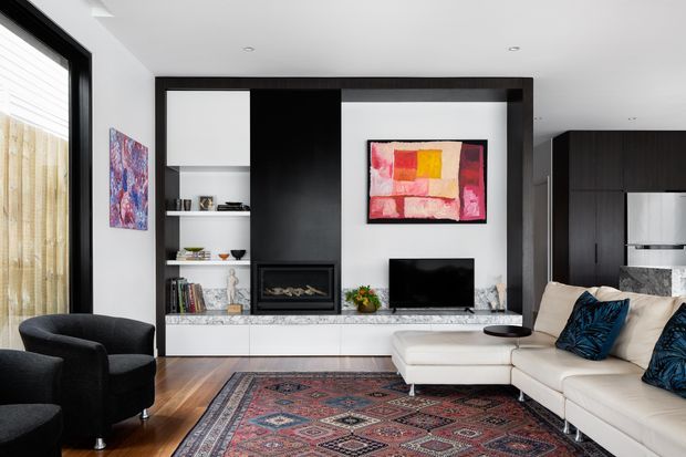 A contemporary open-plan living room with a large white sectional sofa, black armchairs, a patterned area rug, and a built-in dark timber entertainment wall. The joinery includes open shelving, a gas fireplace, low white cabinetry, and a television, with colourful abstract artwork above. The kitchen is partially visible in the background, while large glazing with sheer curtains brings in natural light.