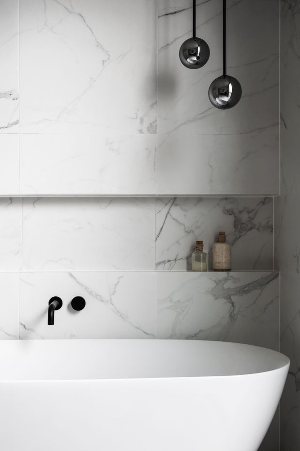A minimalist bathroom featuring a freestanding white bathtub against full-height white marble-look wall tiles with grey veining. A recessed shelf holds two small bottles, while matte black wall-mounted tapware and two black spherical pendant lights provide contrast. The composition is clean, monochromatic, and refined.
