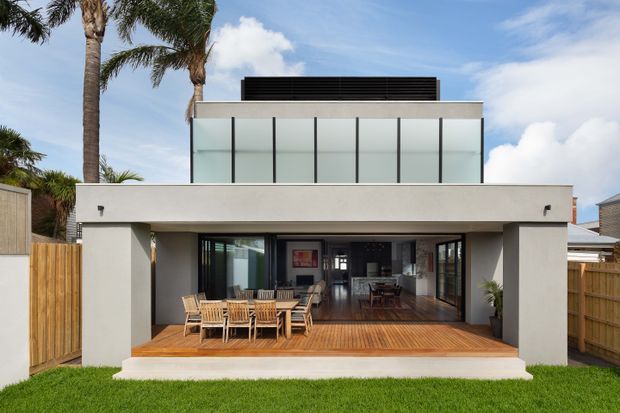 A contemporary two-storey residential home viewed from the rear, featuring a broad flat-roofed form, white rendered walls, a dark upper roof element, and large translucent or frosted upper-level glazing. The ground floor opens onto a timber deck with outdoor dining furniture, while timber boundary fencing, palm trees, and lawn surround the house.