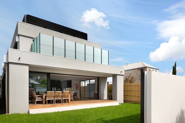 Contemporary multi-level residential home viewed from the rear, with a broad covered outdoor entertaining area, timber dining furniture, large glazed openings, an upper-level terrace with translucent privacy glazing, and clean white rendered walls. A manicured lawn and timber privacy fencing frame the house under a bright blue sky.