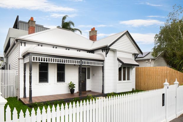 Front exterior view of a renovated single-storey white weatherboard house with a pitched gabled roof, two brick chimneys, a covered timber veranda, decorative black posts and brackets, and a white picket fence. A timber privacy fence and neighbouring buildings are visible, with palm and leafy vegetation in the background.
