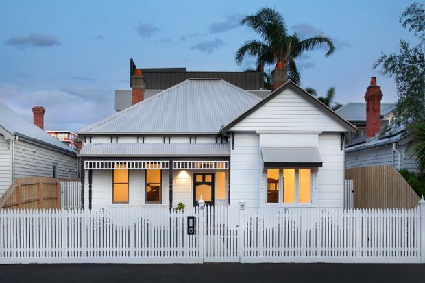 Front exterior view of a compact single-storey house with white weatherboard cladding, a corrugated metal gabled roof, brick chimneys, enclosed front porch, striped awnings, and a white picket fence. The home is set in a dense suburban streetscape with neighboring houses and palm trees visible behind it.
