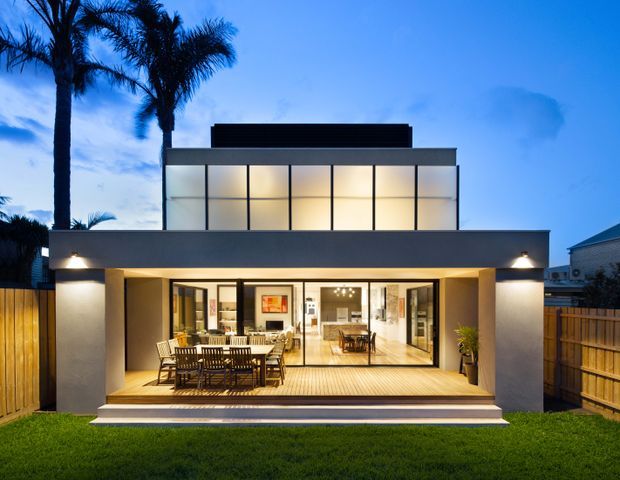 A dusk-time rear exterior view of a contemporary two-storey residence with a broad, open-plan ground-floor living area opening onto a timber deck. The upper level features a large grid of illuminated translucent or glazed panels, while the lower facade is rendered in dark grey with recessed lighting. Outdoor dining furniture sits beneath the covered deck, with timber fencing and tall palm trees framing the property.
