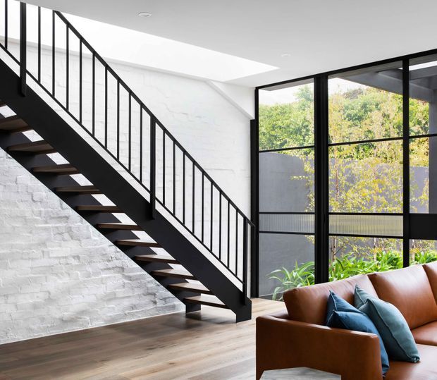 Bright contemporary interior featuring an open-riser staircase with black-painted steel stringers and slender vertical balustrade, set against a white-painted exposed brick wall. Large black-framed floor-to-ceiling glazing opens the living area to greenery outside. A cognac leather sofa with blue cushions occupies the foreground, and wide timber flooring runs throughout the space. A linear skylight or clerestory brings additional daylight above the stair.