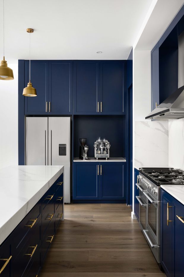 A refined, compact kitchen with deep navy-blue full-height cabinetry, brass pulls, white veined stone benchtops and splashback, stainless-steel appliances, and warm timber flooring. A large island occupies the foreground, while integrated refrigeration, coffee station cabinetry, rangehood and freestanding range define the main work areas. Brass pendant lights and carefully detailed panelled joinery create a polished, high-contrast interior.