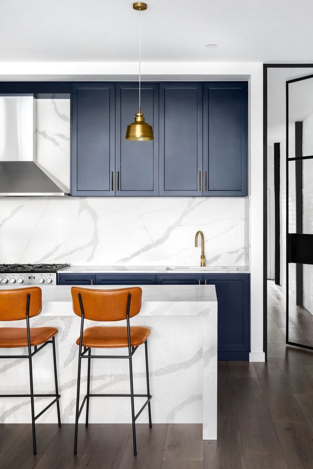 A refined kitchen with deep navy-blue shaker cabinetry, white marble-look slab backsplash and waterfall island, brass pendant lighting and faucet, stainless-steel range, and two tan leather counter stools. Dark timber flooring and black-framed glazed doors add contrast, while the compact layout has a polished, high-contrast contemporary feel.