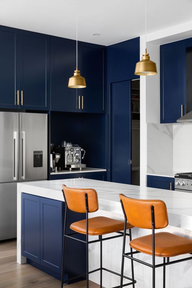 A refined kitchen featuring deep navy-blue shaker-style cabinetry, a large white stone island with waterfall edges, stainless-steel appliances, and integrated coffee station cabinetry. Two cognac leather counter stools with black metal frames sit at the island, while brass pendant lights add warmth. The space is bright and high-contrast, with light timber flooring and white walls and splashback surfaces.