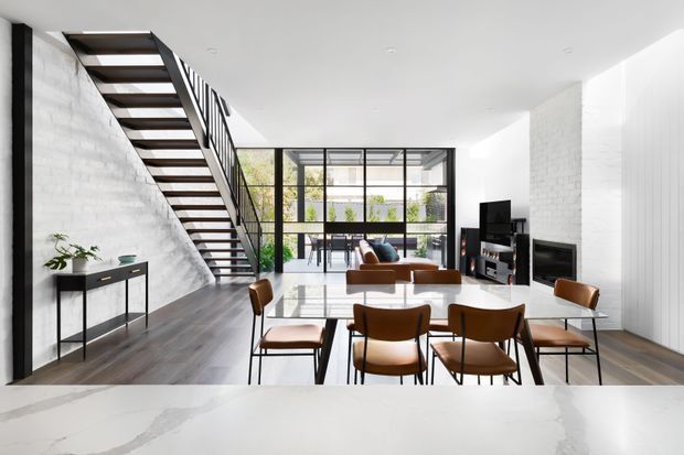 Bright open-plan contemporary interior with a dining area in the foreground, a living room beyond, and a dramatic open-riser staircase along a textured white masonry wall. Full-height black-framed glazing opens the space to greenery, while dark timber flooring, warm brown leather dining chairs, minimalist black joinery, and a fireplace create a refined modern setting. A pale stone or engineered-stone benchtop is visible at the bottom edge.