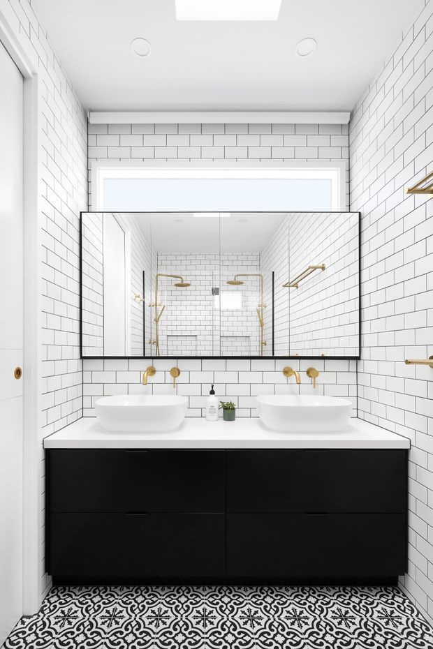 Bright contemporary bathroom with white subway-tiled walls, a large black-framed mirrored cabinet, double white vessel basins on a white benchtop, and a floating matte-black vanity. Brass-gold tapware and shower fittings provide contrast, while patterned black-and-white floor tiles add a decorative feature.