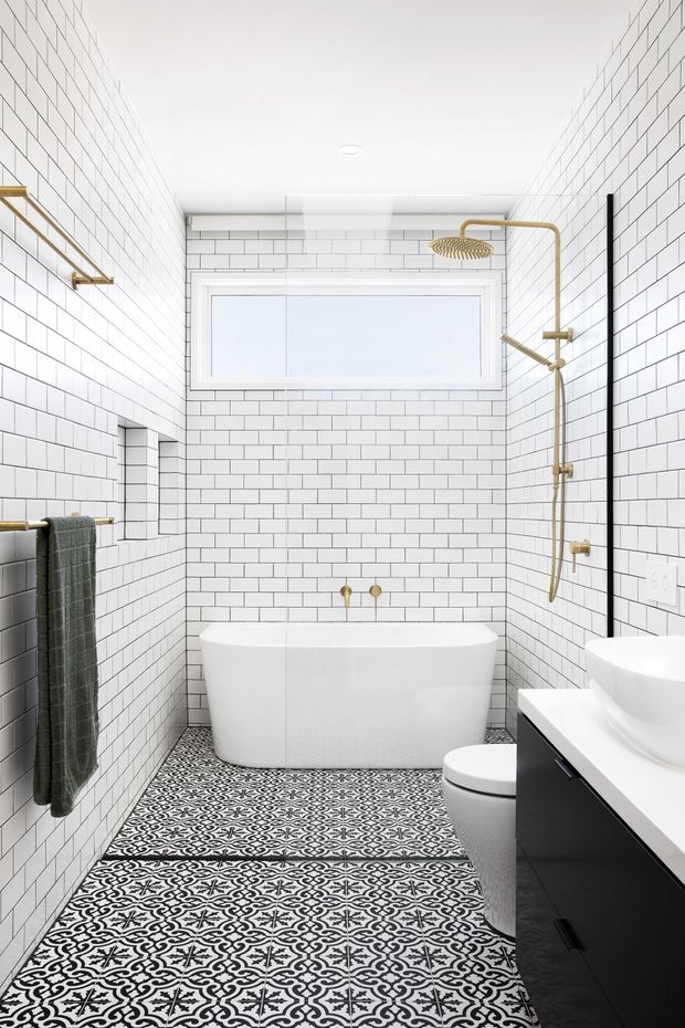 Bright, compact bathroom with white subway-tiled walls, a freestanding white bathtub beneath a horizontal frosted window, patterned black-and-white floor tiles, and a black floating vanity with a white basin. Brushed brass shower fittings, towel rail, and tapware provide warm metallic accents, while a dark towel and black-framed shower screen add contrast.
