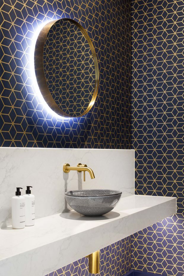 A compact, dramatically styled bathroom vanity featuring dark navy-blue geometric wall tiles with gold linear detailing, a large round backlit mirror with a gold-toned frame, a white marble-look benchtop and integrated shelf, a grey vessel basin, and a brushed gold wall-mounted tap. The space has a luxurious, bold, art-deco-inspired feel with strong geometric patterning and high contrast.