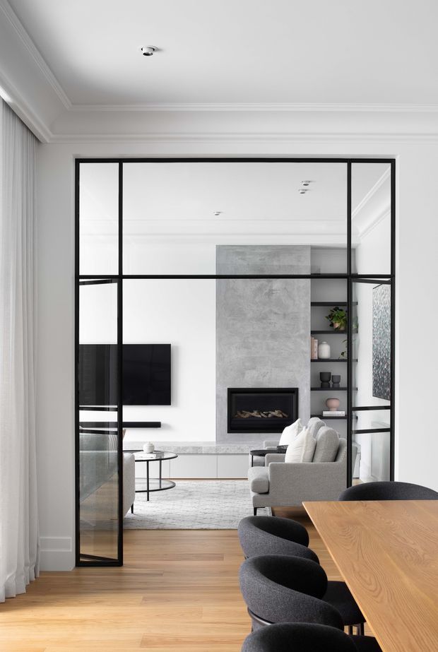 A refined contemporary open-plan interior viewed through a black-framed glazed partition. The foreground contains a timber dining table with dark upholstered chairs, while the adjoining living room features a pale sofa, wall-mounted television, concrete-look fireplace surround, built-in shelving, and low cabinetry. White walls, ornate cornices, sheer curtains, and warm timber flooring create a bright, sophisticated setting.