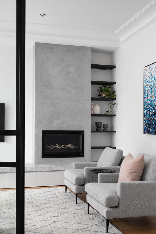 Contemporary living room featuring a built-in linear fireplace set within a textured grey concrete-look feature wall, flanked by open black shelving. The room has white walls with decorative ceiling cornices, pale grey upholstered armchairs, a blush cushion, a patterned neutral rug, black-framed glazing or partitioning, and restrained decorative styling.