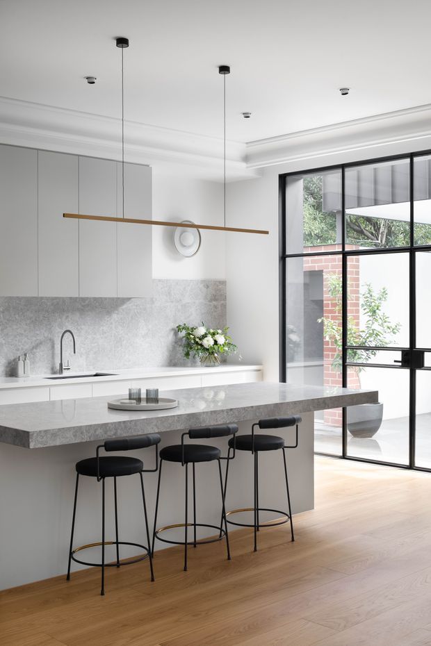Bright contemporary kitchen with a large grey stone island and integrated seating for four. The space features pale grey full-height cabinetry, a matching stone splashback and benchtop, warm timber flooring, black-framed steel-style glazing, and a minimalist linear pendant light. White walls and decorative ceiling cornices add a refined architectural detail, while a courtyard garden is visible beyond the glazing.