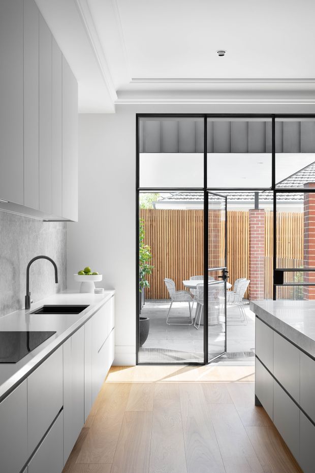 Bright contemporary kitchen with white built-in cabinetry, a dark stone benchtop and integrated sink, pale timber flooring, and a central island. Black-framed glazed doors open directly to a paved outdoor dining courtyard with timber screening and exposed brickwork. The room has a layered decorative ceiling with recessed detailing and abundant natural light.