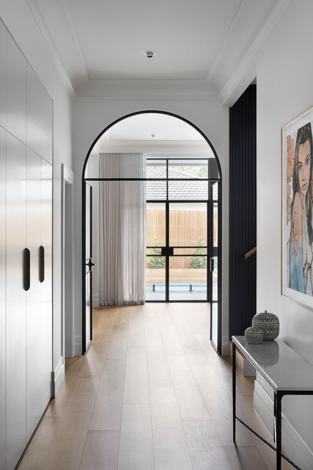 A bright, refined residential hallway or entry passage with pale timber flooring, white built-in cabinetry, and a dramatic black arched steel-framed opening leading toward a glazed outdoor-facing area. Black vertical wall panelling and matching framed glazing add contrast, while a slim console with decorative objects and framed artwork lines the right wall. The space has a high, coffered ceiling and abundant natural light.