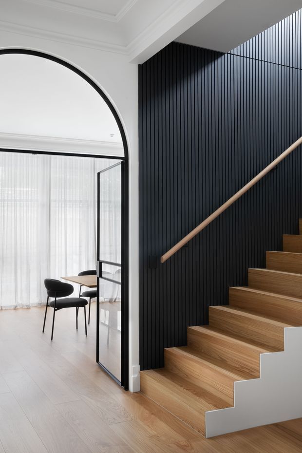 A refined contemporary interior showing a timber staircase rising beside a dramatic black vertically slatted feature wall. The stair has warm natural timber treads, a simple timber handrail, and a white painted side stringer. Beside it is a bright dining area with a small round table and black chairs, enclosed by a tall black-framed glazed door with an arched opening above and sheer curtains. White walls, ornate ceiling cornices, pale timber flooring, and strong black-and-white contrast create a polished modern-classic atmosphere.