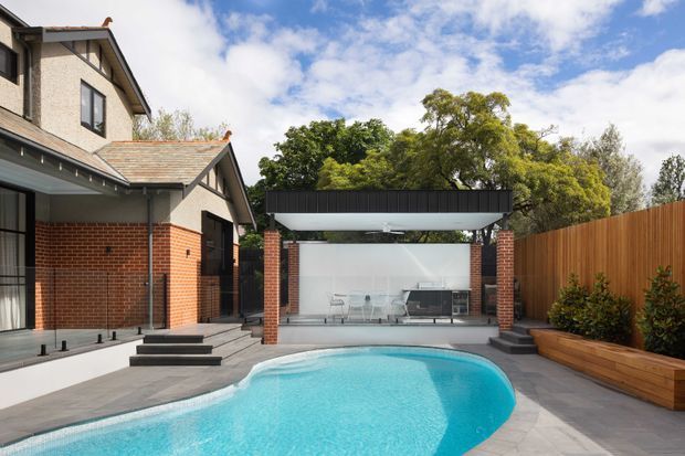 A landscaped residential backyard with a curved swimming pool, paved surrounds, and a covered outdoor entertaining pavilion. The pavilion has a flat black roof, brick supporting columns, a white rear wall, outdoor dining furniture and a built-in barbecue. A brick and rendered multi-level house with tiled pitched roofs borders the pool, while timber privacy fencing and raised timber planters line the garden edge.