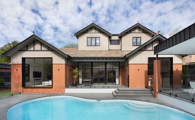 A large multi-wing residential house viewed from the pool area. The facade combines red brick lower walls with pale rendered upper gables, dark timber or metal roof edges, broad black-framed glazing, and a covered entry or terrace at right. A curved swimming pool occupies the foreground, with steps leading up to the central glazed living area. The architecture has a contemporary interpretation of traditional gabled forms.