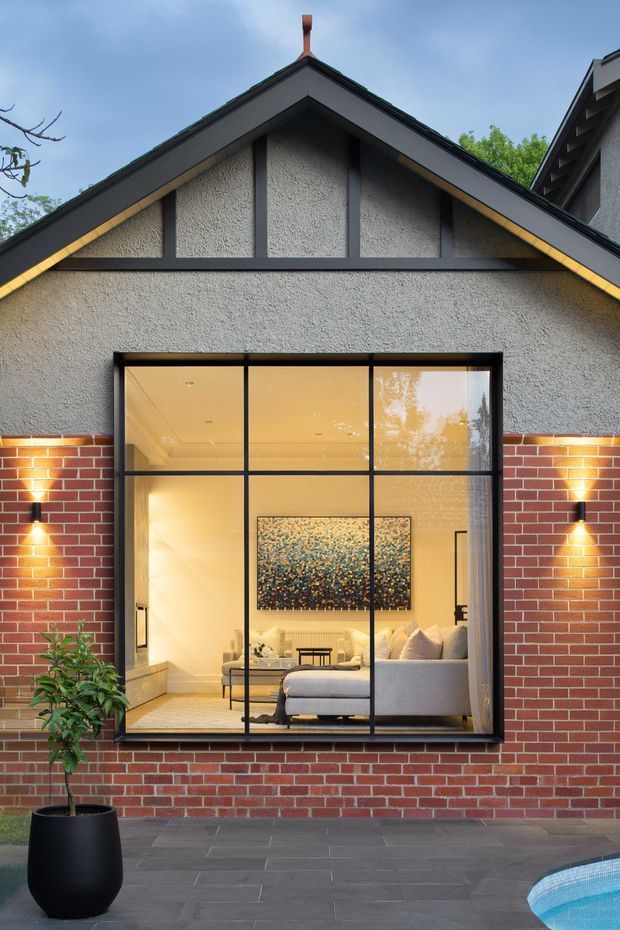 Exterior view of a gabled residential facade with textured grey render above red brickwork, dark timber or metal trim, and a large black-framed window revealing a warmly lit living room. A paved terrace, potted plant, and the edge of a swimming pool are visible in the foreground.