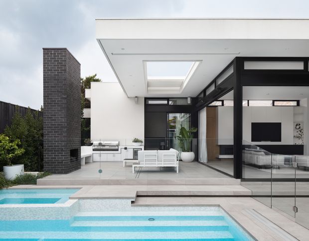 Contemporary residential courtyard exterior showing a white flat-roof pavilion opening onto a tiled terrace and swimming pool. Black-framed floor-to-ceiling glazing connects the outdoor seating area with a modern living room, while a skylight brings light into the covered terrace. A dark brick feature wall and outdoor barbecue area add contrast.
