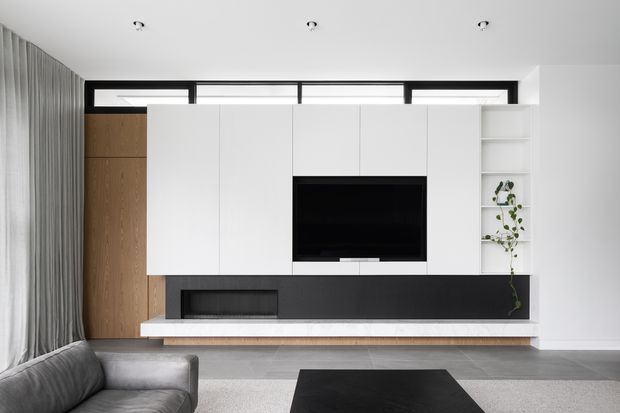 Contemporary living room featuring a wall-to-wall built-in media unit with matte white cabinetry, a recessed black television niche, open shelving with a trailing plant, and a dark feature base. Warm timber wall panelling, full-height grey curtains, a low grey sofa, and a black coffee table create a restrained modern interior. High horizontal windows sit above the joinery, and recessed ceiling lights provide subtle illumination.