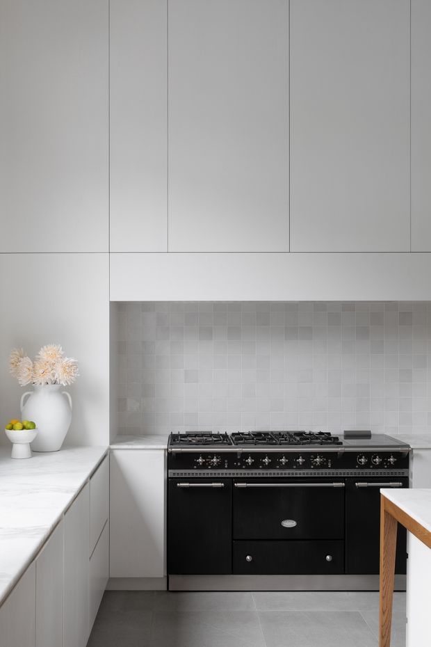 A refined contemporary kitchen with full-height pale grey cabinetry, a large black range cooker, light grey tiled splashback, white marble-look benchtops, and grey tiled flooring. Minimal styling includes a white vase with dried flowers and a small bowl of fruit.