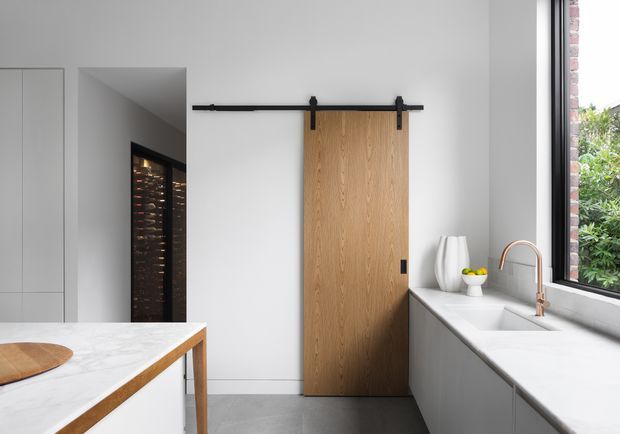 Contemporary kitchen interior with white cabinetry and a pale stone island and benchtop. A warm timber barn-style sliding door with black hardware contrasts against the white walls, while a large window above the sink brings in natural light and views of greenery. A dark wine storage area is visible through the opening at left.