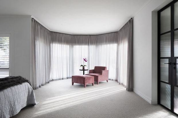 Bright, refined bedroom with a curved bay of floor-to-ceiling sheer curtains, pale carpet, and a small mauve upholstered seating arrangement beside a bed. A black-framed glazed door and plantation-shuttered window add strong contrast to the light interior.