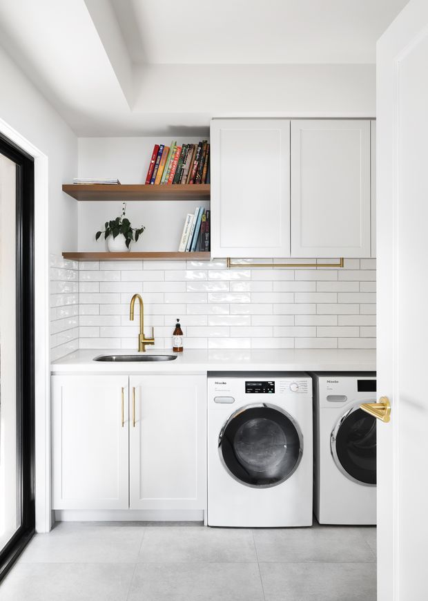Bright, compact laundry room with white raised-panel cabinetry, a white glossy subway-tile splashback, grey tiled flooring, open timber shelving with books and a plant, a stainless sink, and gold-toned tapware and handles. Two front-loading white washing machines are positioned beneath the benchtop. The room is viewed through an open doorway and has black-framed glazing at left.