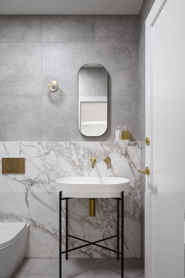 Compact, refined bathroom featuring large grey concrete-look wall tiles above veined white marble-look wall cladding, a white vessel basin on a slim black metal stand, brushed gold tapware and flush plate, an arched rounded-corner mirror, and a white door. The partial freestanding bathtub reinforces the bathroom setting.