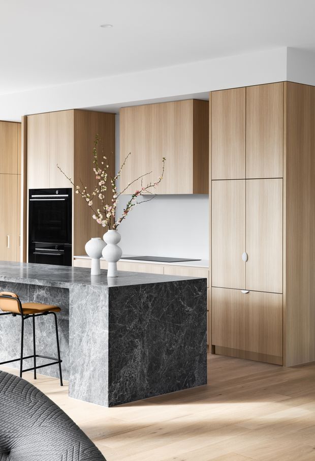 A bright contemporary kitchen with full-height light timber cabinetry, integrated black oven, pale benchtops, and a dramatic dark veined stone island with waterfall sides. A pair of sculptural white vases with flowering branches sits on the island, with a timber-and-black bar stool nearby. The kitchen opens toward a partially visible living area with a grey upholstered sofa.