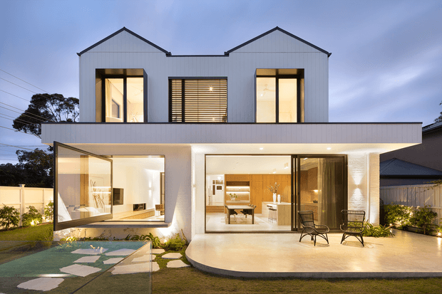 A contemporary two-storey suburban house photographed at dusk. The white facade features two steep gabled upper volumes, large glazed openings, a broad flat-roofed ground-floor terrace, and warm interior lighting. The outdoor terrace has dark-framed lounge chairs, while stepping stones cross the landscaped lawn toward the home.
