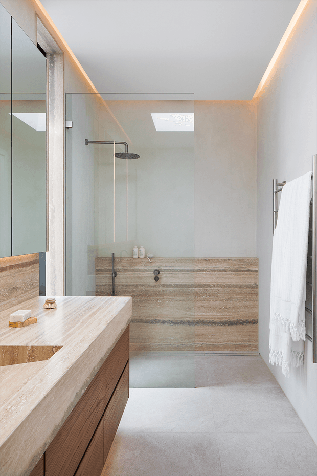 Bright contemporary bathroom with a walk-in glass shower, skylight, warm concealed perimeter lighting, a large travertine-look stone vanity and shower wall, timber vanity cabinetry, and a heated towel rail. The bathroom has a calm, refined, natural material palette.