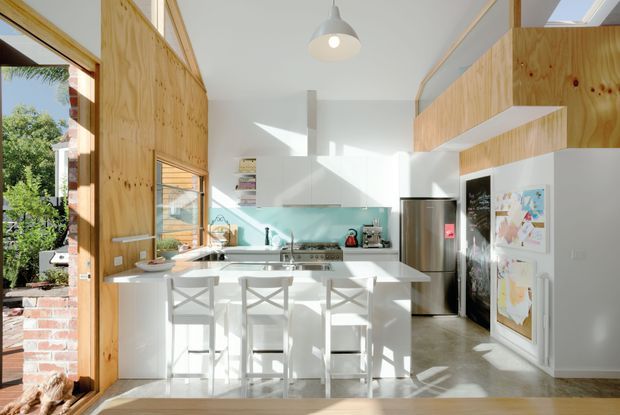 Bright, open-plan contemporary kitchen with white walls and cabinetry, a pale timber-lined feature wall and overhead storage, a turquoise glass or tile splashback, stainless-steel appliances, and a white island with three cross-back stools. Large openings bring in abundant daylight and provide views to greenery and brickwork outside.