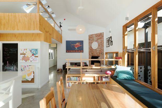 Bright open-plan living and dining area with a double-height white ceiling, exposed timber mezzanine and balustrade, extensive timber-framed glazing, polished concrete flooring, a large timber dining table, and built-in window seating. A brick feature wall, open shelving, books, artwork, and colourful cushions add warmth and character. A skylight and clerestory-style glazing bring in abundant natural light.