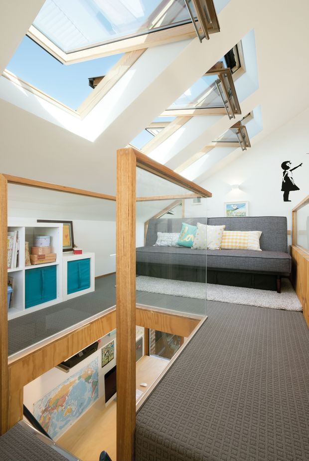 Bright multi-level interior centered on a compact living or sitting area beside an open stairwell. The space features a dark grey sofa with patterned cushions, grey carpet, white walls, extensive timber balustrades and posts, built-in white shelving with turquoise cabinet fronts, and a glass stair guard. A series of large overhead skylights and roof windows floods the double-height circulation space with natural light. A black wall silhouette graphic and framed artwork provide small decorative accents.