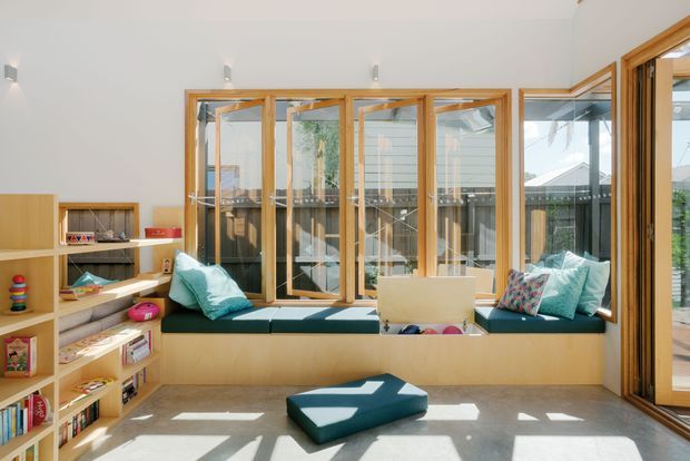 Bright, sunlit family living space with a built-in window seat running along a wall of timber-framed glazing. Light timber shelving and joinery provide open storage for books and toys, while teal cushions and a low upholstered ottoman add colour. The room has a clean, contemporary feel with strong indoor-outdoor visual connection through the extensive windows and glazed door.