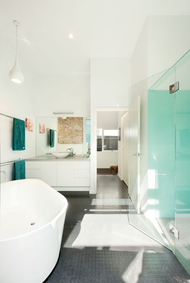 Bright contemporary bathroom with a freestanding white bathtub, wall-mounted white vanity with vessel sink, large mirror, turquoise glass shower enclosure, and dark textured rubber flooring. A doorway leads to an adjacent space, while strong sunlight creates crisp shadows across the room.