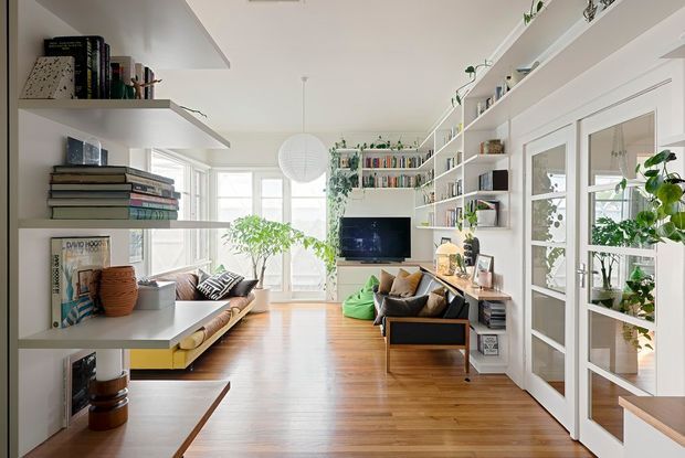 Bright, narrow open-plan living room with extensive white built-in shelving and open display storage along both sides. The room features warm timber flooring, a built-in low sofa with colourful cushions, a black-framed lounge chair, a wall-mounted television, indoor plants, books, and a large round pendant light. Glazed white-framed French doors open to a plant-filled adjoining space, while abundant windows bring in natural light.