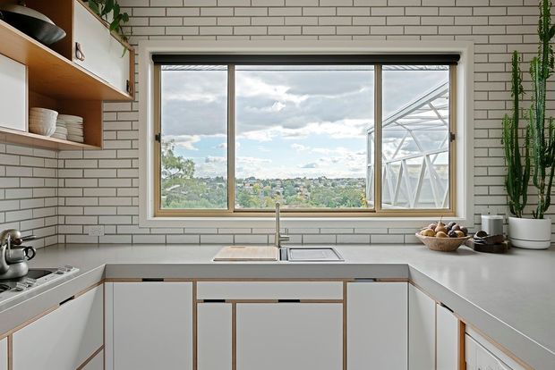 Bright contemporary kitchen with a U-shaped white cabinetry layout, pale benchtops, and a large horizontal window overlooking a leafy suburban landscape. White brick-pattern tiled walls, timber open shelving, integrated stainless-steel sink and tapware, and indoor greenery create a clean, warm modern interior.