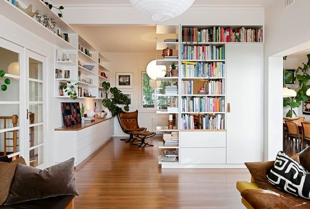 Bright, refined open-plan living and study space with extensive white built-in shelving and cabinetry, a large floor-to-ceiling bookcase, warm timber flooring, glass-panel French doors, indoor plants, and leather lounge seating. A reading chair sits near the central shelving, while a dining area is visible through the opening at right. The room has decorative ceiling moulding and layered pendant lighting, creating an inviting, book-filled home interior.