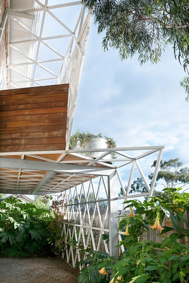 Exterior view of a lightweight, elevated architectural structure with white-painted steel trusses, extensive open framing, and warm horizontal timber wall and soffit cladding. The structure is integrated into a lush garden with dense tropical and subtropical planting, gravel paths, and abundant daylight. The image emphasizes the building's exposed structural framework and indoor-outdoor connection.
