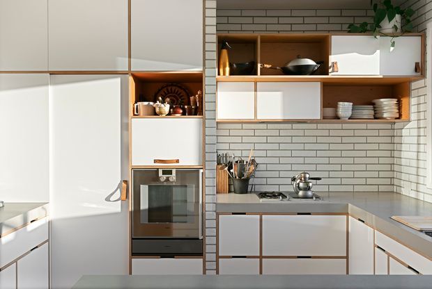 A bright, compact contemporary kitchen with extensive white flat-panel cabinetry outlined by warm timber trim. The room features a built-in oven, a tiled benchtop wall with open timber shelving, stainless steel cooking appliances, and a pale grey worktop. White subway tiles cover the backsplash and side wall, while ceramics, cookware, and a trailing plant add restrained visual detail. The cabinetry and shelving create a clean, highly organised built-in composition.