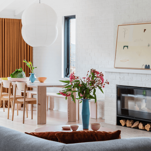 Bright contemporary living and dining area with a white-painted brick fireplace wall, black-framed window, timber vertical screen, pale timber dining table and chairs, sculptural pendant light, and a large blue vase with leafy branches. A framed abstract artwork and wood-burning fireplace add warmth and character.