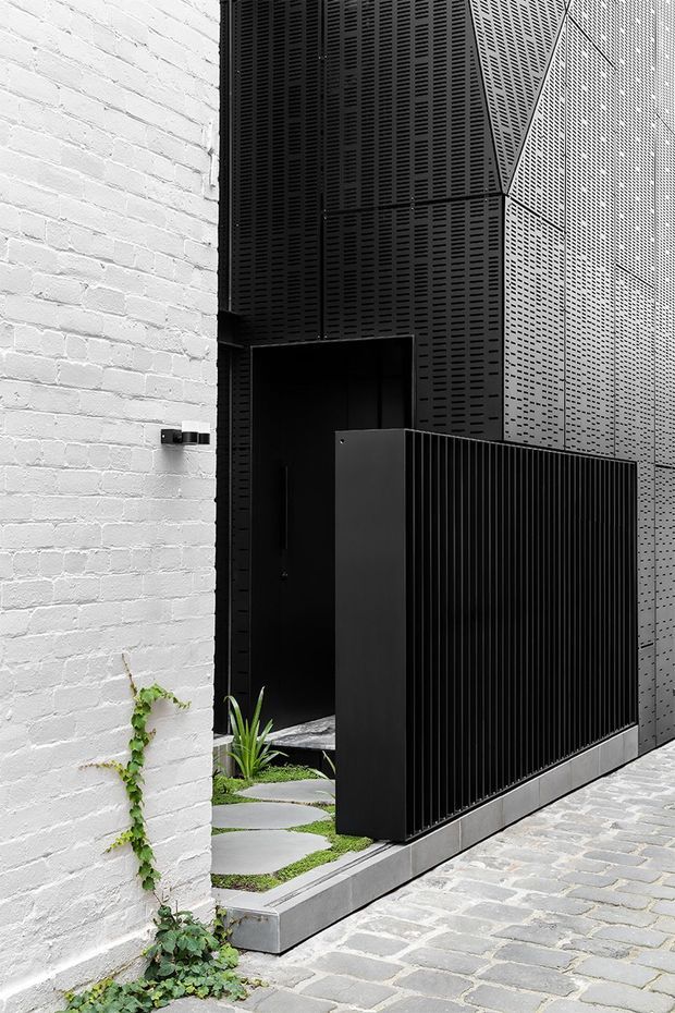 A narrow contemporary architectural entrance with a white-painted brick wall contrasting against a tall black perforated metal facade and matching vertical-slat privacy gate. Grey stone pavers lead past a small planted entry garden with stepping stones and low greenery. The composition is monochromatic, graphic, and strongly defined by the contrast between white masonry and black metalwork.